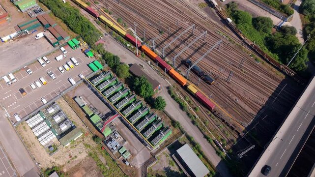 Aerial view tracking over a complex arrangement of commercial power generation machinery enclosures, exhaust systems, and service lines inside a rail maintenance yard.