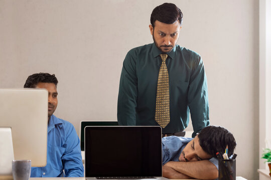 An angry boss behind two corporate employees with one of them sleeping at workdesk.