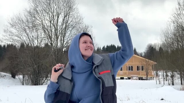 Smiling youth in blue winter jacket raises fist outdoors in snowy yard under overcast sky, with leafless trees and rustic cabin creating cheerful seasonal atmosphere