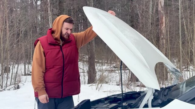 Man in Red Puffer Vest and Brown Hoodie Holds Large Satellite Dish beside Open Car Hood in Snowy Winter Forest Outdoor Scene, Focused Expression, Rugged Winter Work Setup