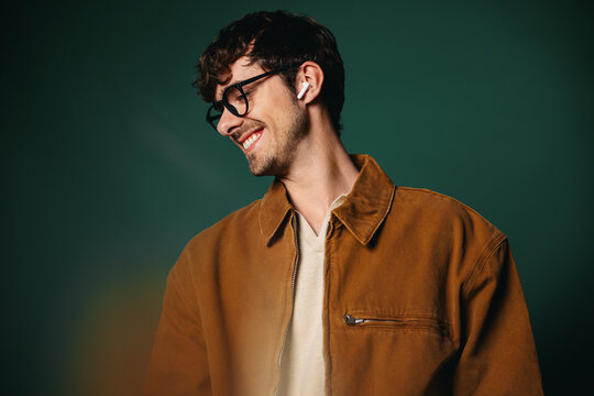 Smiling man with glasses wears earbuds in a green studio portrait