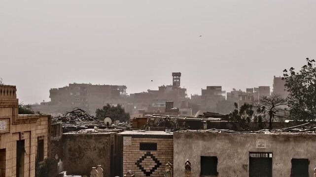 Dusty panoramic view over Cairo City of the Dead necropolis, Egypt, historic Islamic mausoleum domes rising above palm trees and crumbling brick tomb buildings in sandstorm haze.
