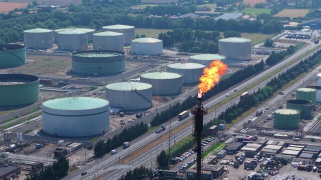 Tight close-up aerial shot tracking the thermal combustion flames of a refinery flare stack against a backdrop of industrial storage silos and processing units.