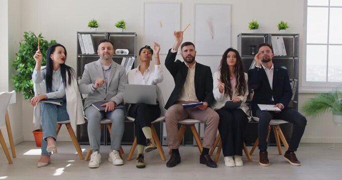 Women and men audience sitting on chairs in row, raising hands, wanting to ask coach questions, taking part in seminar or training session, posing against office interior. Business people voting