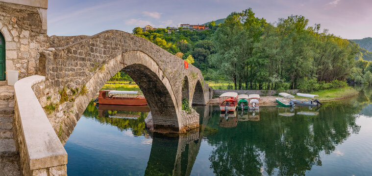 Iconic Rijeka Crnojevica river with an ancient stone bridge, boats, and lush green mountainous landscape reflecting in still water in Montenegro