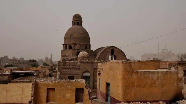 Moody view over Cairo necropolis, Egypt, Mamluk era mausoleum domes and crumbling tomb structures among palm trees with sandstorm casting orange haze over city.