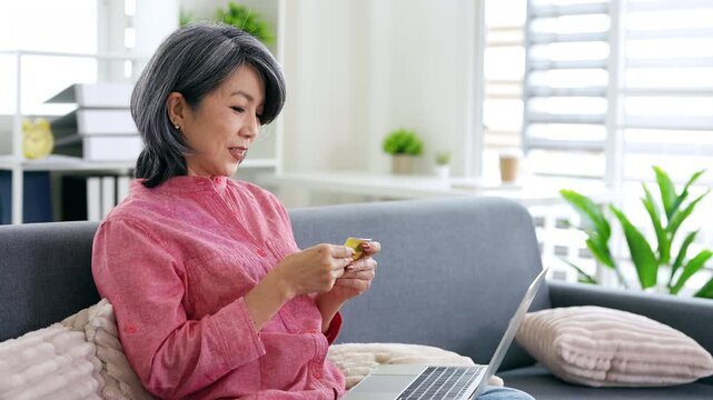 Online  shopping. Happy mature woman with credit card and laptop sitting on sofa, confirming internet order.
