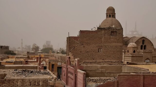 Historic City of the Dead cemetery in Cairo, Egypt, Mamluk mausoleum domes visible through palm trees above old brick tomb structures in dusty hazy weather.