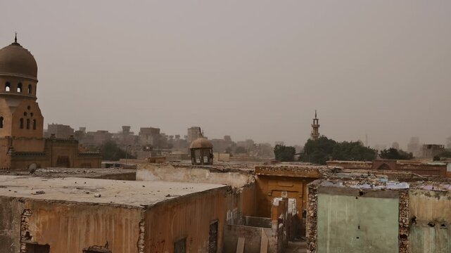 Moody view over Cairo necropolis, Egypt, Mamluk era mausoleum domes and crumbling tomb structures among palm trees with sandstorm casting orange haze over city.