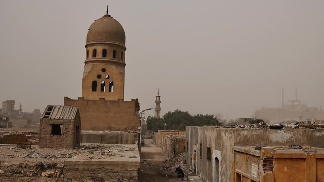 Medieval Islamic mausoleums and tomb structures of Cairo City of the Dead necropolis, Egypt, surrounded by palm trees and old buildings under orange dusty sky.