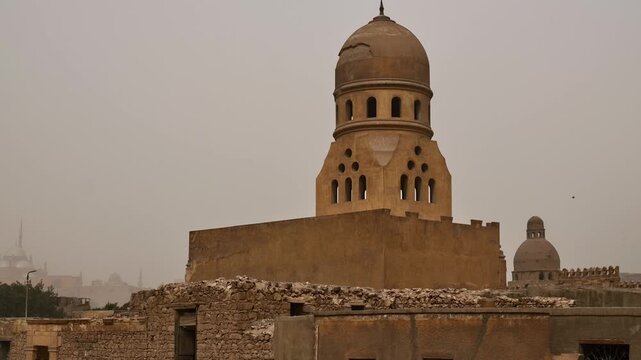 Historic City of the Dead cemetery in Cairo, Egypt, Mamluk mausoleum domes visible through palm trees above old brick tomb structures in dusty hazy weather.
