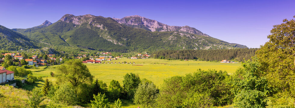 Rural village nestled in a green valley, surrounded by lush forests and the towering Lovcen mountain range under a clear sky