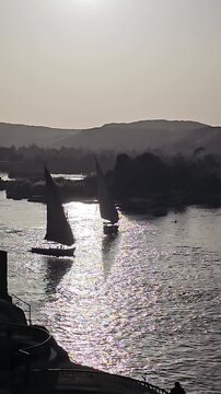 Stunning golden hour on Nile River at Aswan, Egypt, multiple traditional felucca sailboats gliding on shimmering water with silhouetted desert hills at sunset.