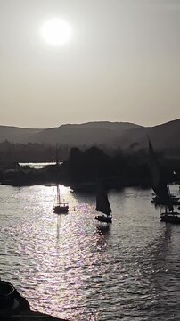 Golden sunset over Nile River at Aswan, Egypt, traditional felucca sailboats with glowing sails silhouetted against shimmering water, desert hills and islands in warm hazy light.