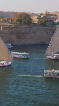 Busy Nile waterfront at Aswan, Egypt, white sailed feluccas and colorful tourist ferries on blue water with Nubian village houses and desert horizon beyond.