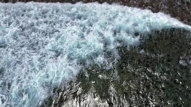 Drone looks straight down as waves roll over submerged rocks at Cabo del Sol, then tracks vertically with the second wave to reveal a glimpse of the shoreline at the edge of the frame.