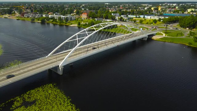Stunning aerial footage showing a modern arch bridge with traffic crossing a wide river, with a boat passing underneath and creating a wake, connecting to a city with lush green banks