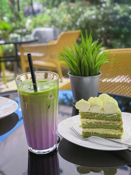 Colorful layered iced matcha Layered matcha drink with berry purple milk and ice stands beside slice of green cake with cream on cafe. Matcha drink and dessert, food menu, bakery, beverage promotion