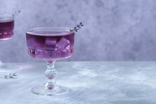 Refreshing purple lavender cocktail served in an elegant vintage coupe glass with ice cubes and a fresh lavender sprig. Minimalistic composition on gray background