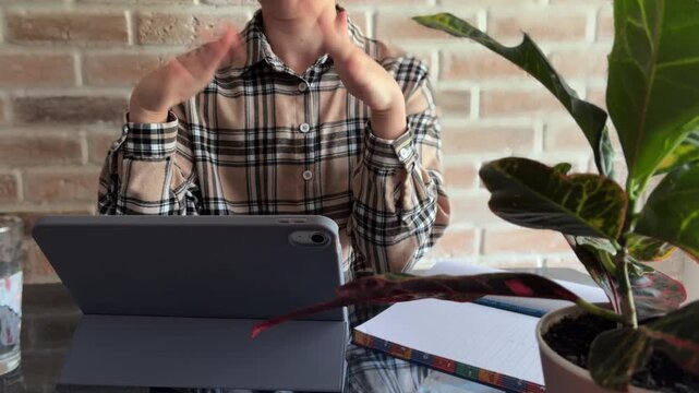 Frustrated young woman gesturing while studying online, experiencing technical difficulties or stress during a video call or exam, sitting at a desk with a tablet in a modern home office