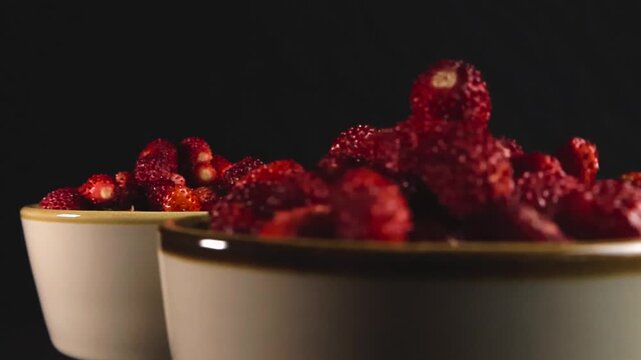 Closeup of two white ceramic bowls brimming with fresh, ripe wild strawberries, slowly gliding past the camera against a dark background, highlighting their vibrant color and texture
