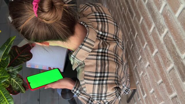 Young student holding a mobile phone with a green screen, studying and browsing at her desk from an overhead perspective, getting ready for an important exam