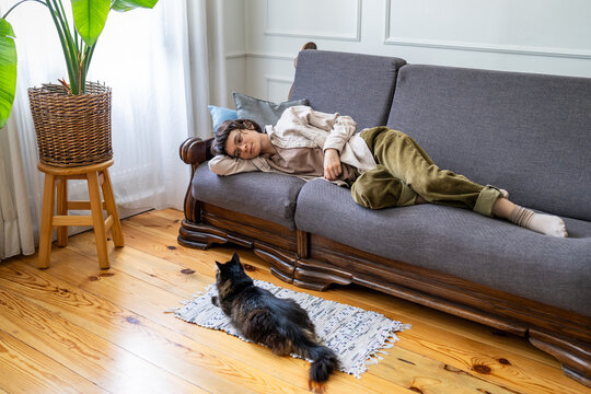Lazy happy woman peacefully relaxing on couch and observing domestic cat resting on rug in room, positive bored female practicing procrastination and self care, spending time in peace and solitude.