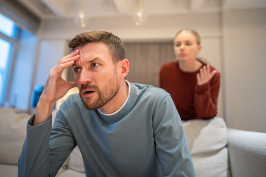 Frustrated angry husband sitting on sofa irritated by wife unstoppable blaming and arguments, married couple problems, mad ignorant male partner turned from complaining woman, complex relationships.