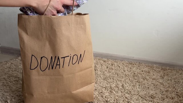 Charitable woman placing used clothes into a paper bag labeled donation, preparing old garments for recycling, supporting a thrift store, and helping people in need