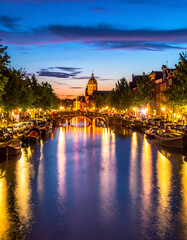 Obraz premium European canal city at twilight, historic architecture and glowing lights reflected on water, vibrant dusk sky over buildings and trees lining the waterway