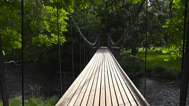 Personal point of view walking along a long wooden suspension bridge over a dark river, surrounded by lush green trees and foliage during a bright and sunny summer day