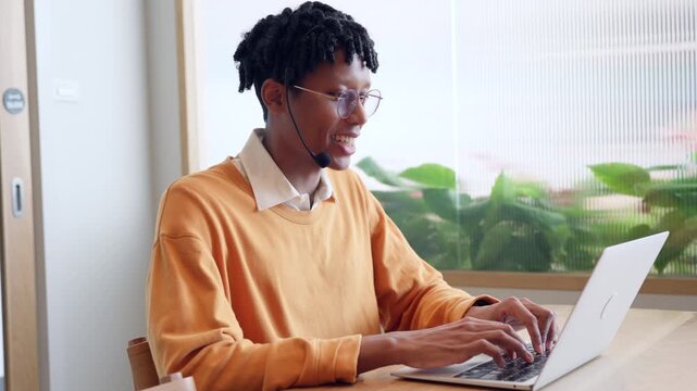 Friendly African American customer support agent with headset typing on laptop at office desk