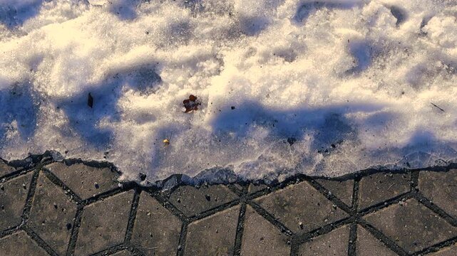 Aerial perspective gliding over a stone pavement sidewalk partially covered with melting snow and ice, with sunlight casting long shadows creating a cold and beautiful winter background