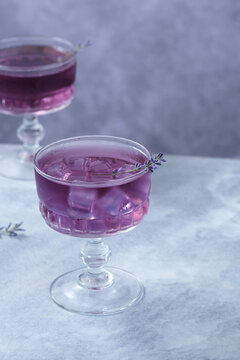 Refreshing purple lavender cocktail served in an elegant vintage coupe glass with ice cubes and a fresh lavender sprig. Minimalistic composition on gray background