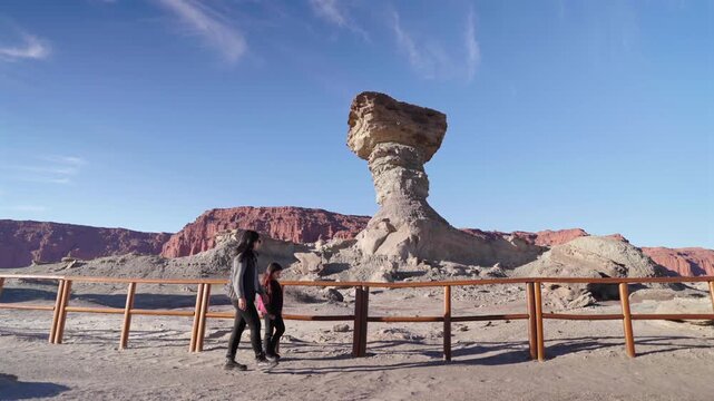 Mother and young daughter walking past the massive El Hongo geological monument on clear sunny afternoon. Slow motion right sliding camera view.
