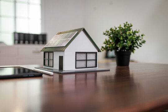 Small house model on a wooden desk with a smartphone and potted plant in a bright modern room, real estate and home planning concept