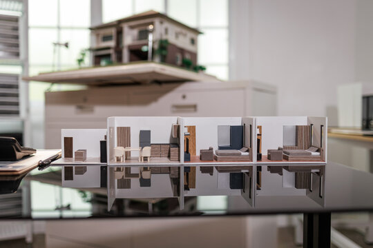 Small architectural model of a modern apartment layout displayed on a glossy table with a soft blurred office background and reflection