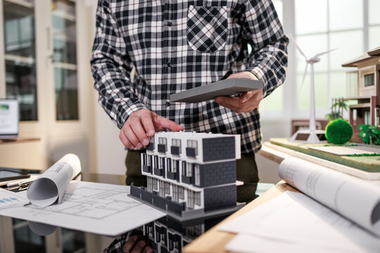 Architect working with a tablet and building model on blueprints in a bright office, urban planning, design review, and construction concept.