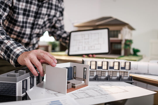 Architect hand arranging a house model with floor plans and tablet in a home design office, real estate planning and construction concept.