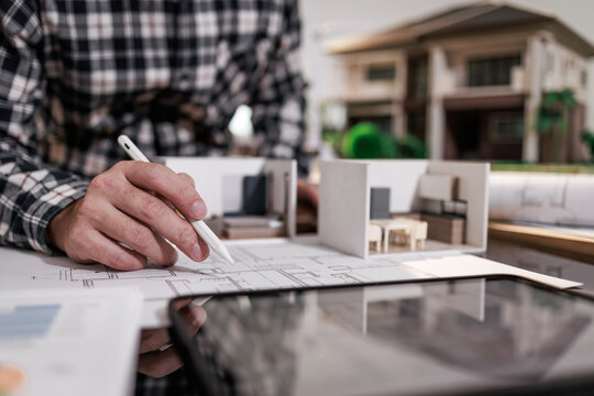 Architect working on house plans with tablet and small model home, planning a residential project with sketch drawings and blurred house in the background