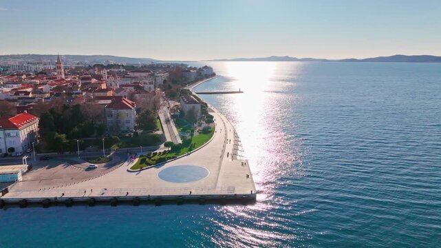 Aerial drone view of Zadar's famous Sea Organ and Greeting to the Sun beside the calm Adriatic Sea. Sunny weather, reflections, old town backdrop and a few visitors strolling.