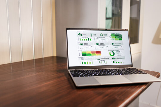 Laptop on a wooden desk displaying a green business dashboard with charts and icons in a bright office.
