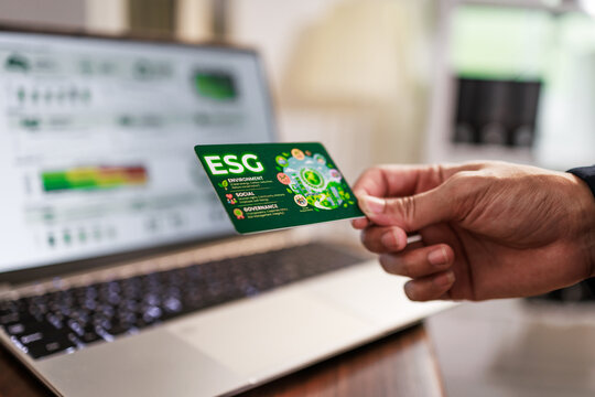 A hand holding a green esg card in front of a laptop with a blurred screen, suggesting sustainable business, finance, and digital work