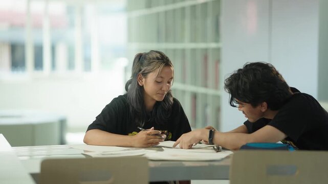 Fashion student pointing at a design sketch while explaining an idea to a classmate seated across a table in a bright university library