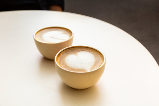 Caf&eacute; cappuccino dans deux tasses beiges avec un latte art motif c&oelig;ur en premier plan et un motif fleur en second plan, servi sur une table de terrasse de caf&eacute;

