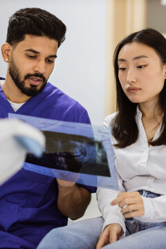 A male doctor in scrubs shows a dental X-ray to a female patient, discussing the results during a consultation