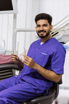 A smiling male dentist in blue scrubs sits in a dental chair holding two metal instruments, ready for a procedure