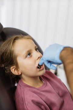 Close up view of hands of professional male doctor in blue uniform and gloves, making examination or curing caries and toothache for his little patient, cute kid girl, sitting in dental chair.