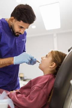 A male dentist wearing blue gloves carefully examines a young girl's teeth with dental instruments in a bright clinic setting
