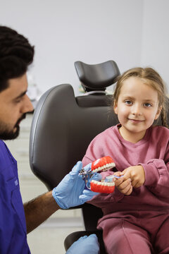 A dentist wearing blue gloves demonstrates proper brushing techniques on a dental model to a young girl sitting in a dental chair
